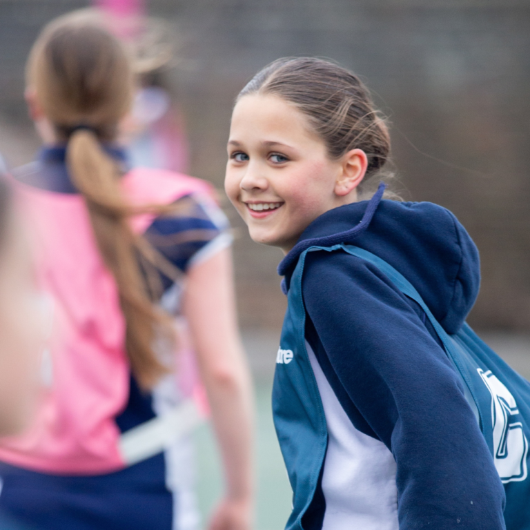 Smiling girl with blue hoodie and backpack on a sports field, among teammates blurred in the background.