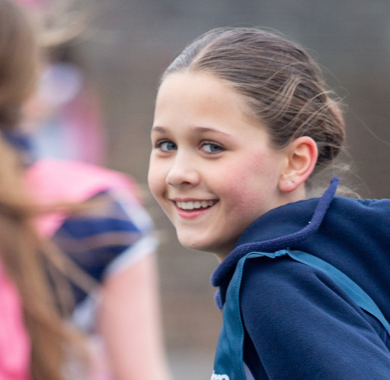 Smiling girl with blue hoodie and backpack on a sports field, among teammates blurred in the background.
