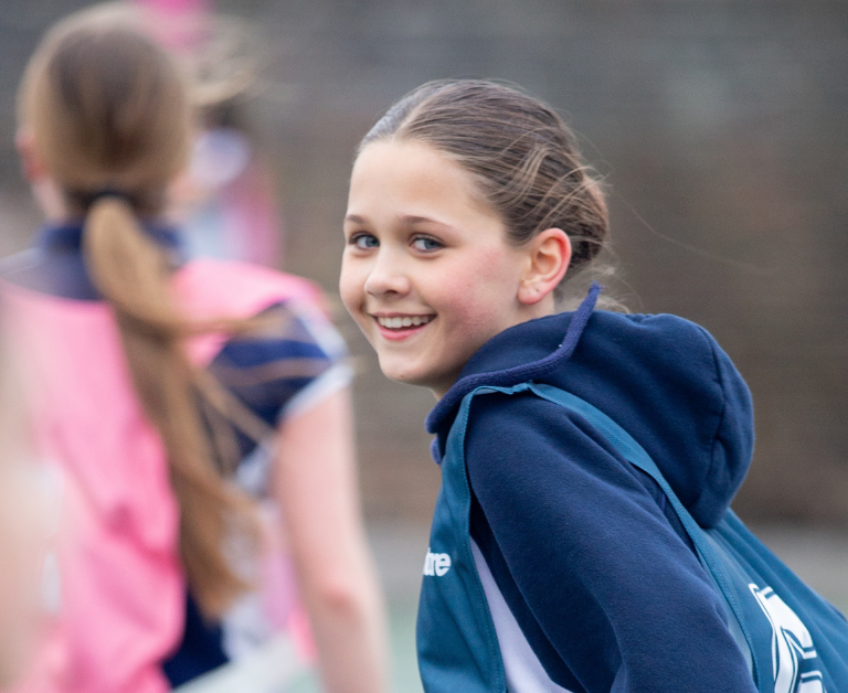 Smiling girl in a blue hoodie with a teal backpack standing with friends outdoors, blurred background.