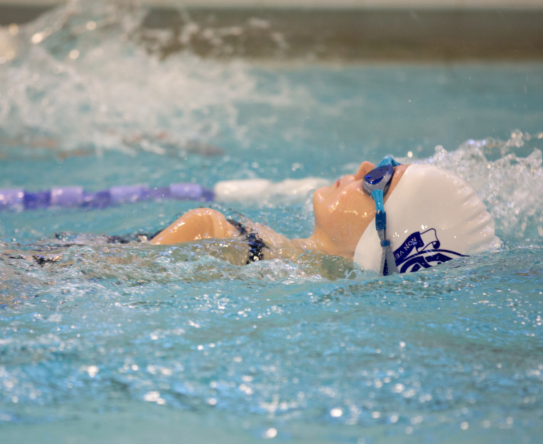 Swimmer in a white cap and blue goggles gliding on their back in a pool lane with splashing water around.